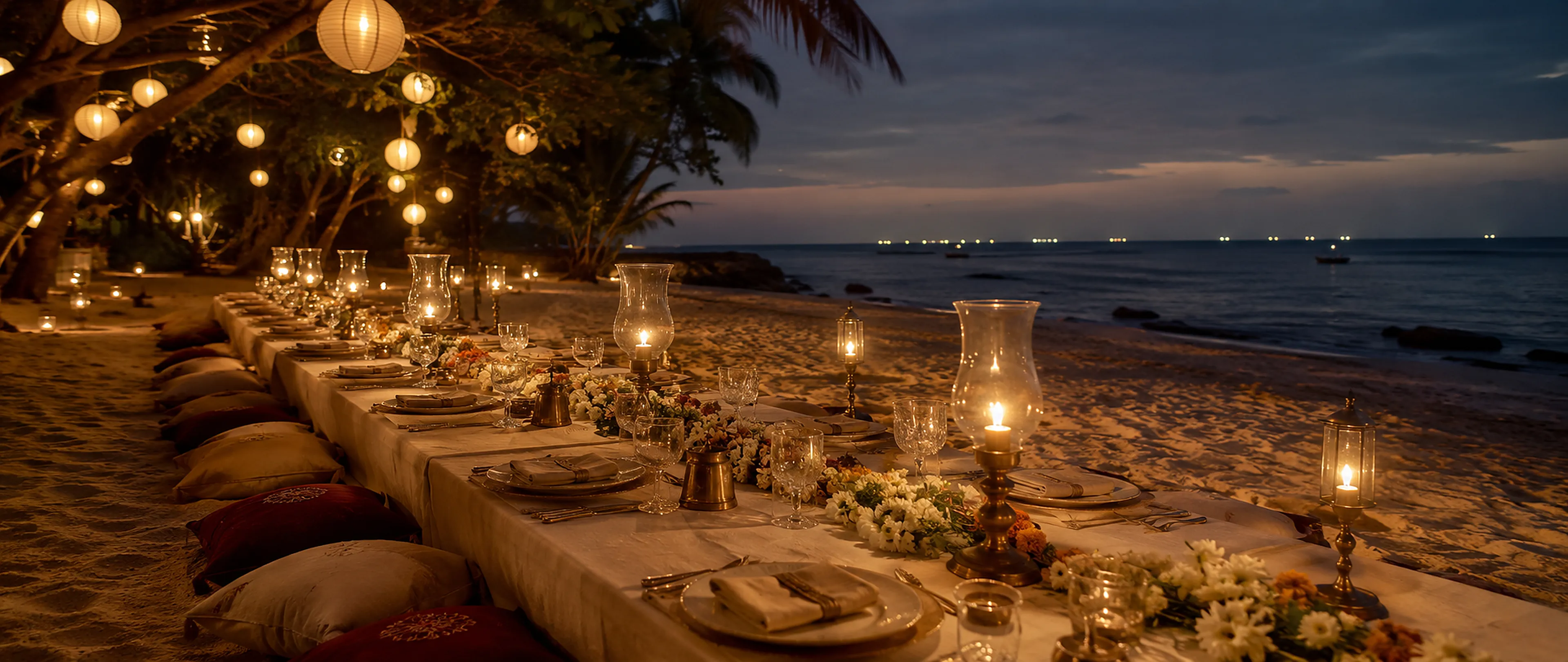Long dinner tables set on a beach at night, lit by paper lanterns